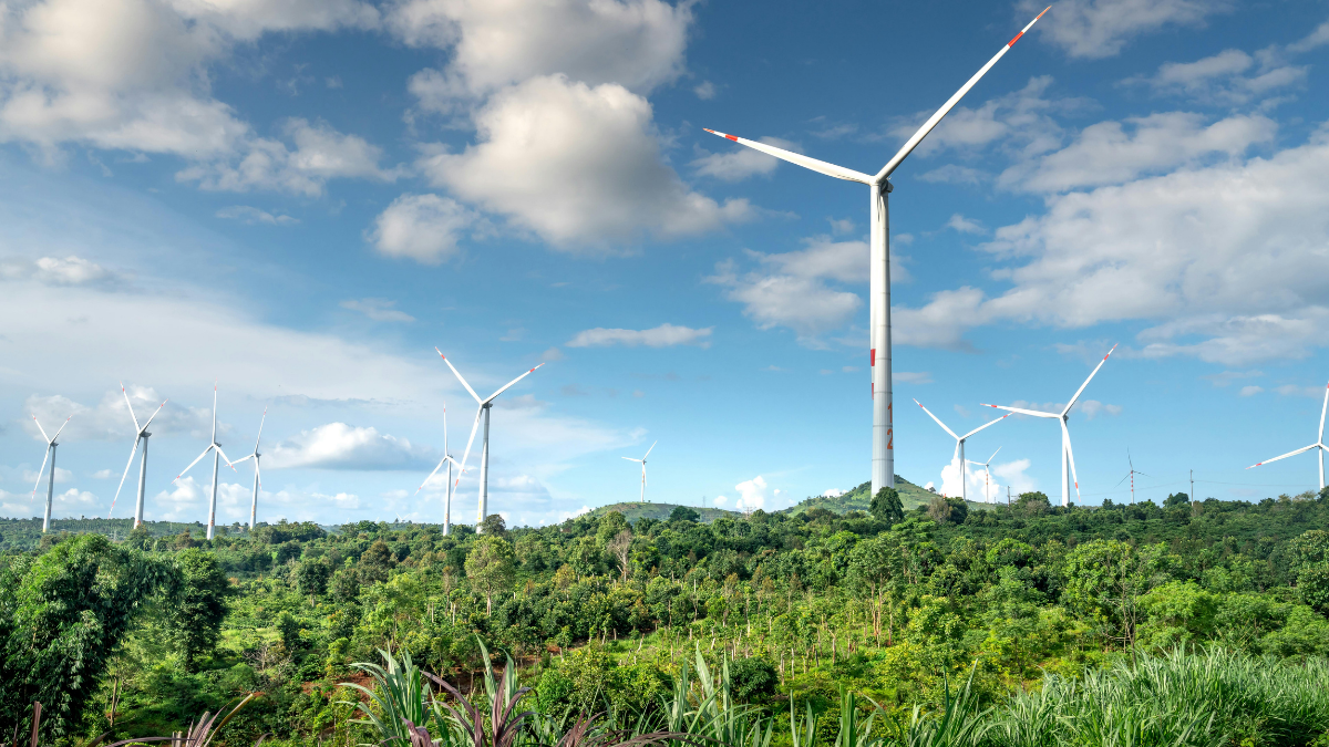 Wind turbines stand on forested hills under a cloudy sky