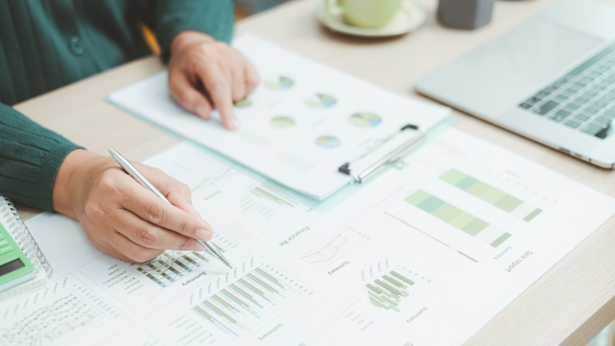 Person pointing with a finger at a chart on a sheet of paper next to a laptop on a table