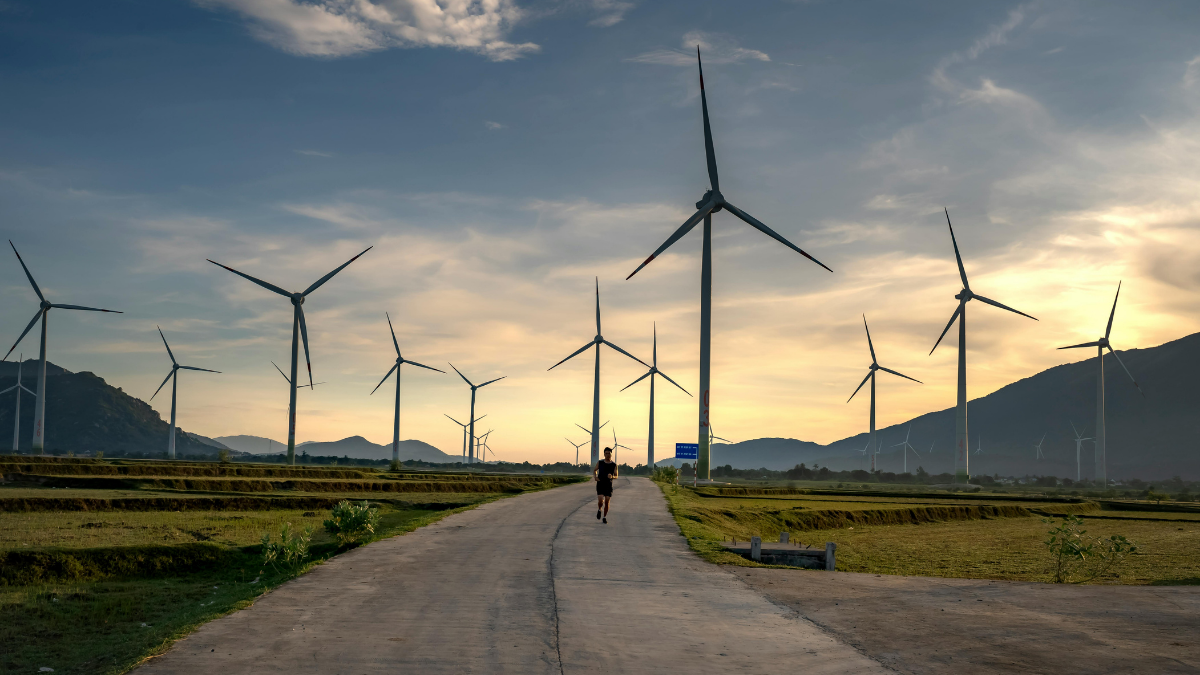 A landscape with wind turbines along a road, a person walking on it, against a cloudy sky and mountains in the background.