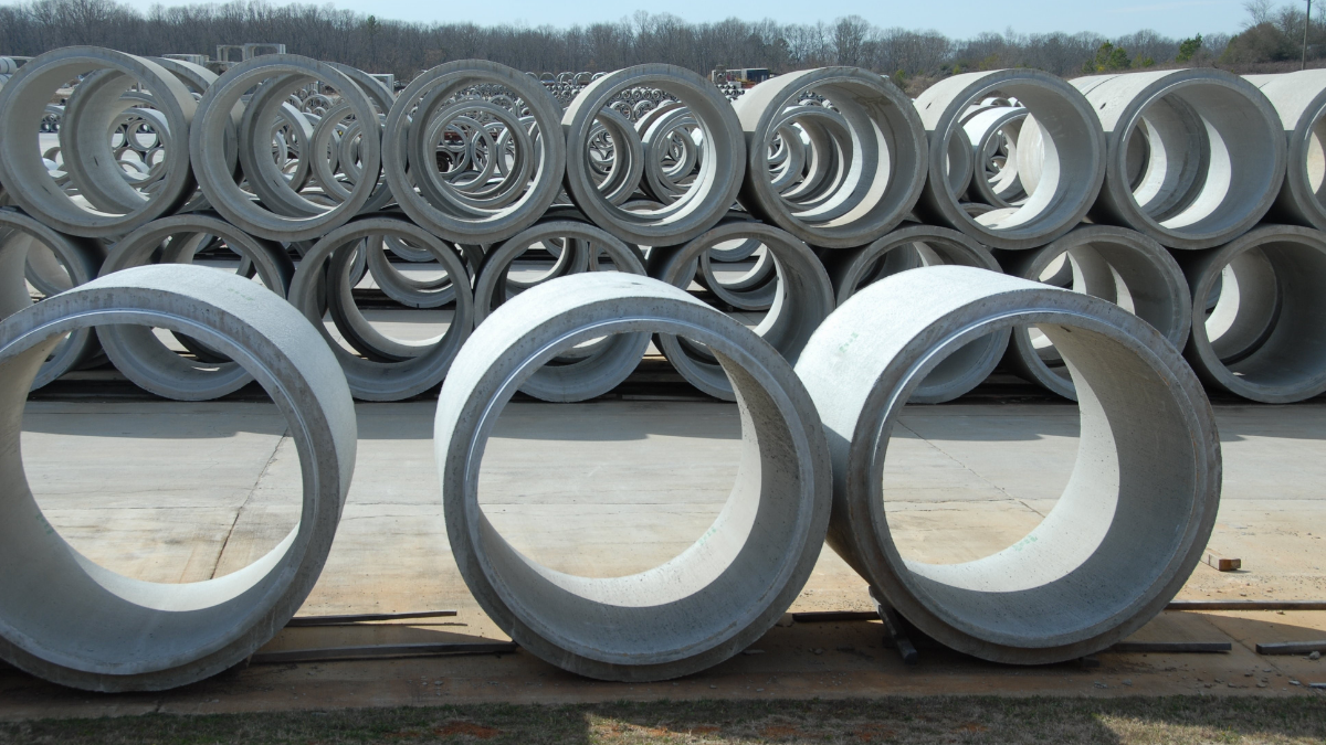 Several large concrete pipes are stacked in rows, both vertically and horizontally, in an open storage yard.