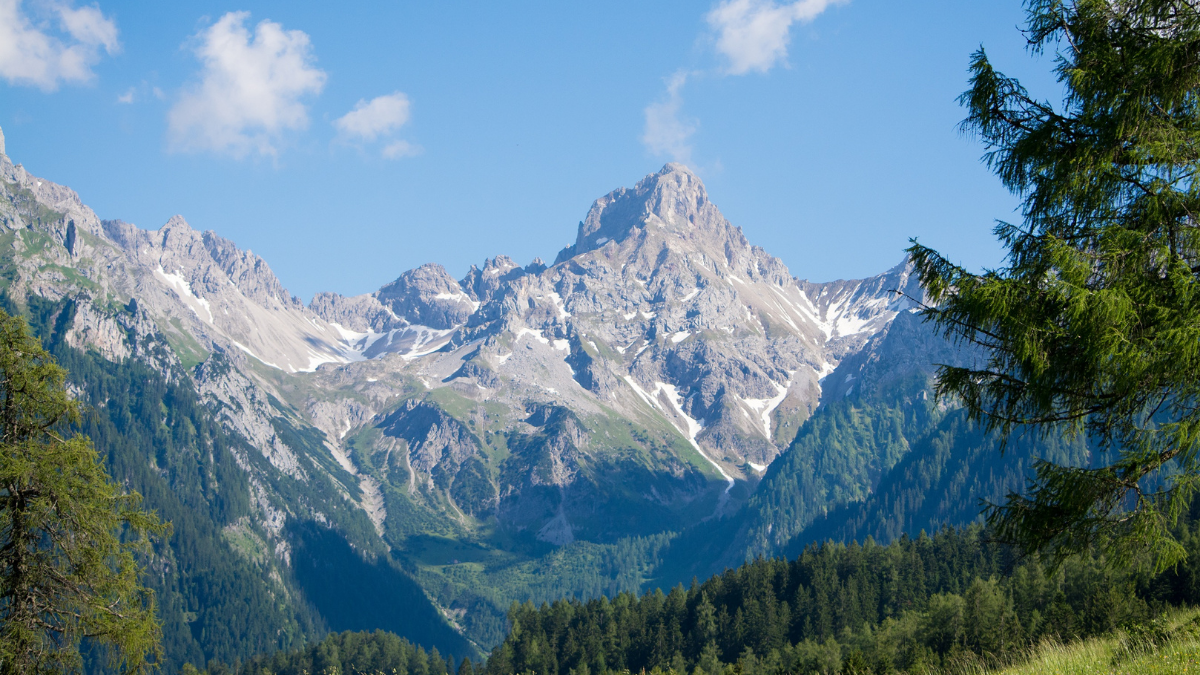 Mountain landscape with snow-covered peaks, forested slopes, and a blue sky with clouds